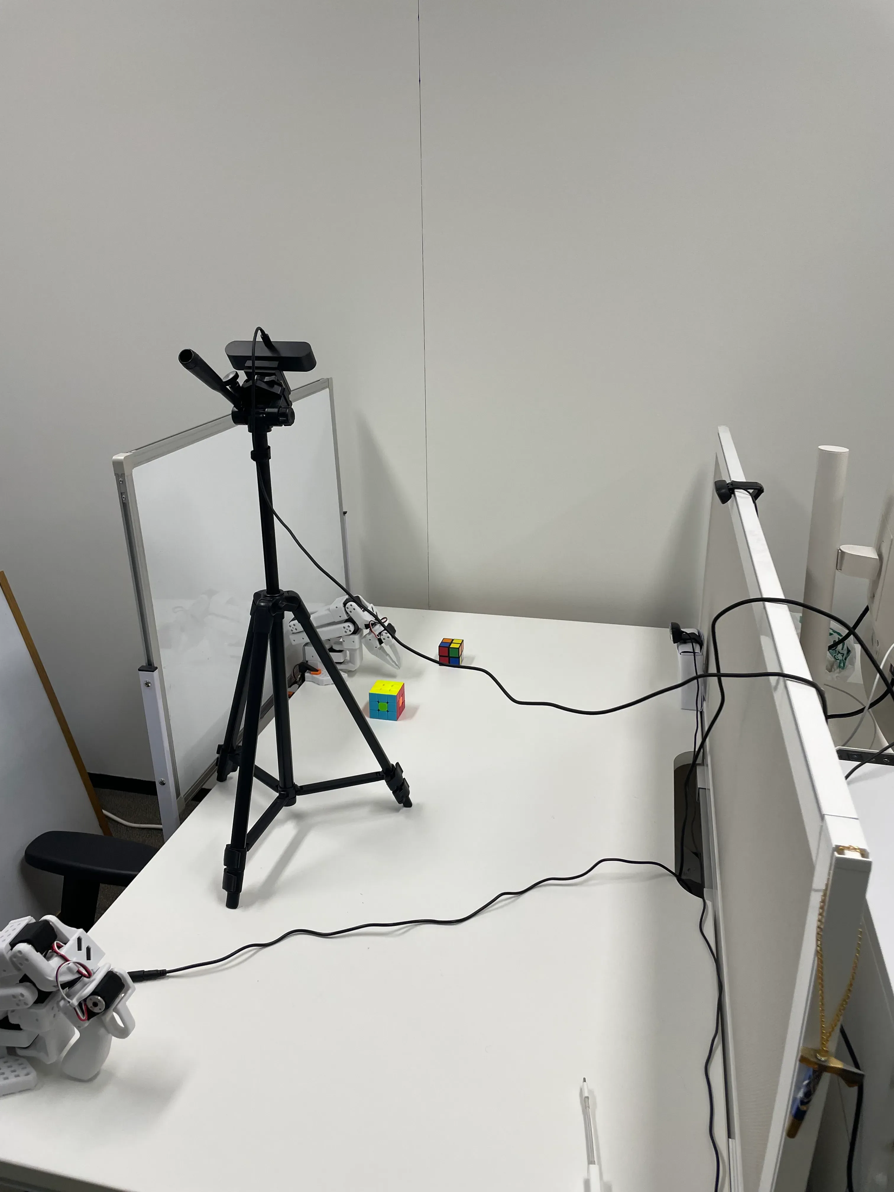 Real-world tabletop experiment setup with SO-101 robot arms, overhead camera tripod, and divider panels.